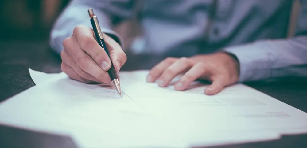 A person writing on a document with a pencil at a desk.